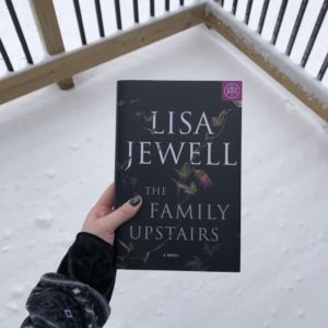 The book Family Upstairs by Lisa Jewell held over a snow covered deck. 